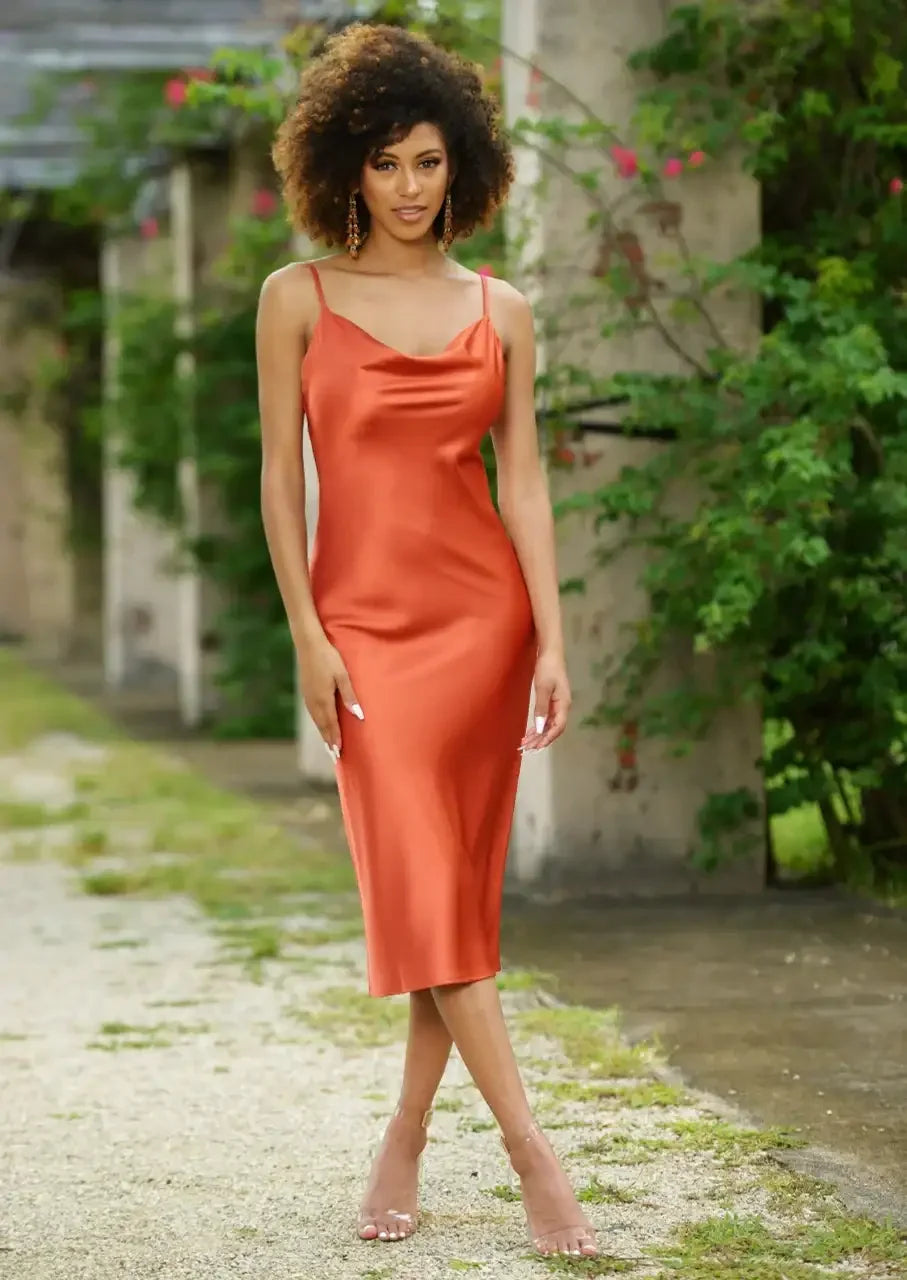 A woman stands confidently wearing a sleek, orange satin dress with thin straps. She poses in a green, overgrown outdoor setting with stone columns and soft, natural lighting.