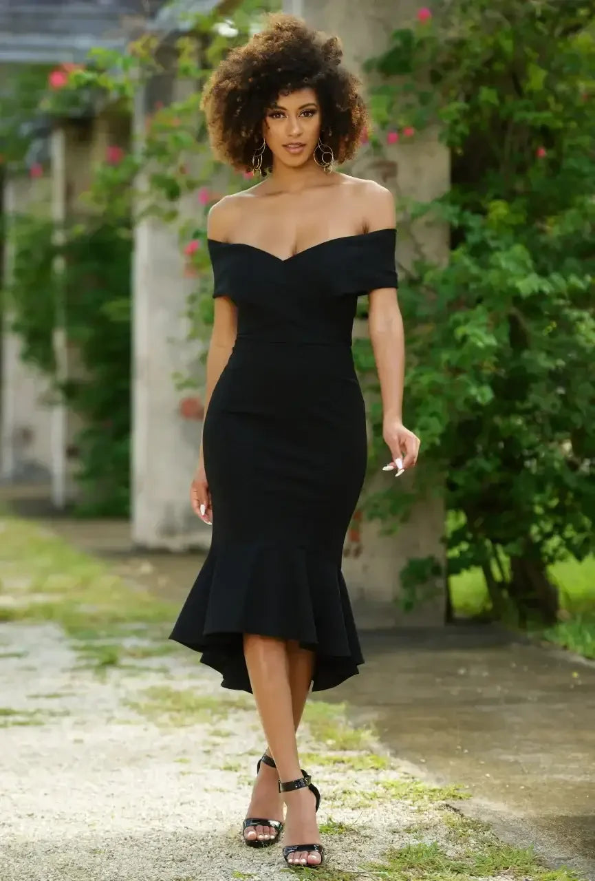 A woman walks gracefully in a fitted black dress with a flared hem. She is surrounded by lush greenery and a textured background, exuding elegance and confidence.