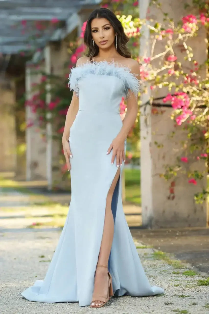 A woman stands in a light blue gown with off-the-shoulder feathers and a thigh-high slit. She poses in an outdoor setting adorned with pink flowers and rustic architecture.