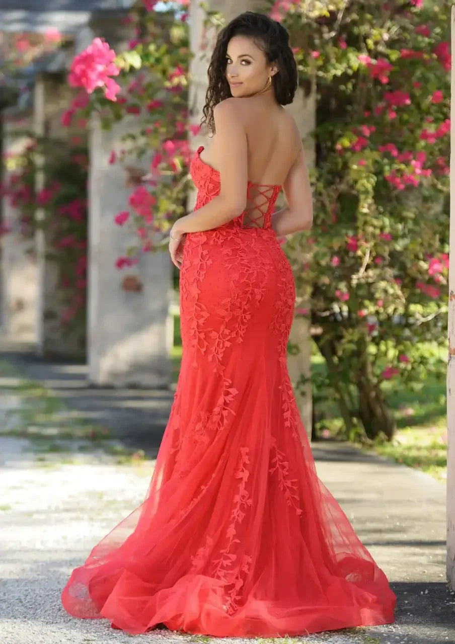 A woman in a fitted, red, lace evening gown poses gracefully outdoors. She stands with her back turned, revealing intricate floral details, surrounded by blooming bougainvillea and stone columns.