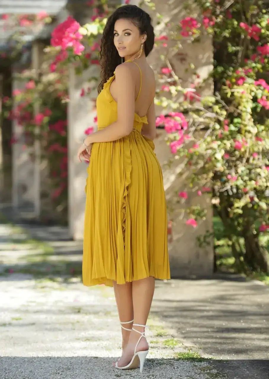 A woman in a bright yellow dress with ruffles stands confidently, turning slightly away from the camera. She is surrounded by blooming pink flowers and an outdoor pathway.