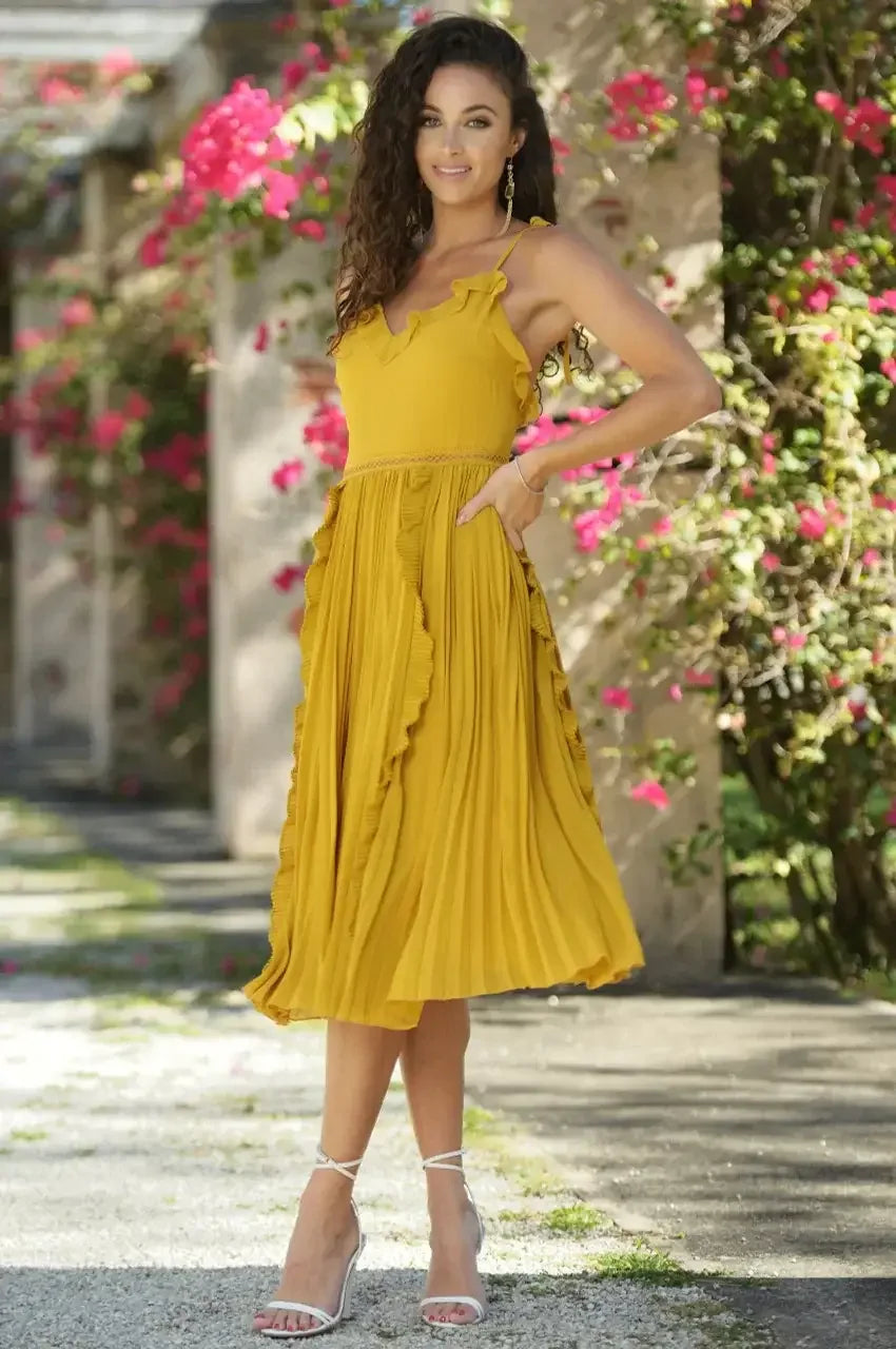 A woman stands gracefully in a vibrant, pleated yellow dress, smiling amidst lush pink flowers and greenery, suggesting a warm, sunny outdoor setting. She wears stylish white heels.