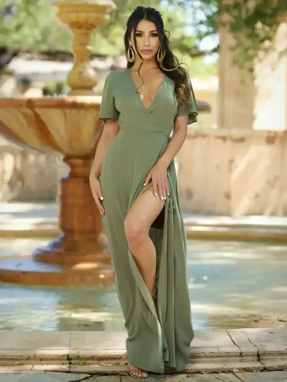 A woman in an olive green dress with a deep V-neck stands by a decorative fountain, showcasing a confident pose against a sunlit outdoor setting with blurred greenery in the background.