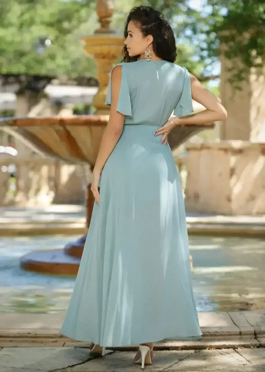 A woman in a light blue, flowing maxi dress poses near a large fountain in a lush outdoor setting, basking in sunlight and surrounded by greenery.