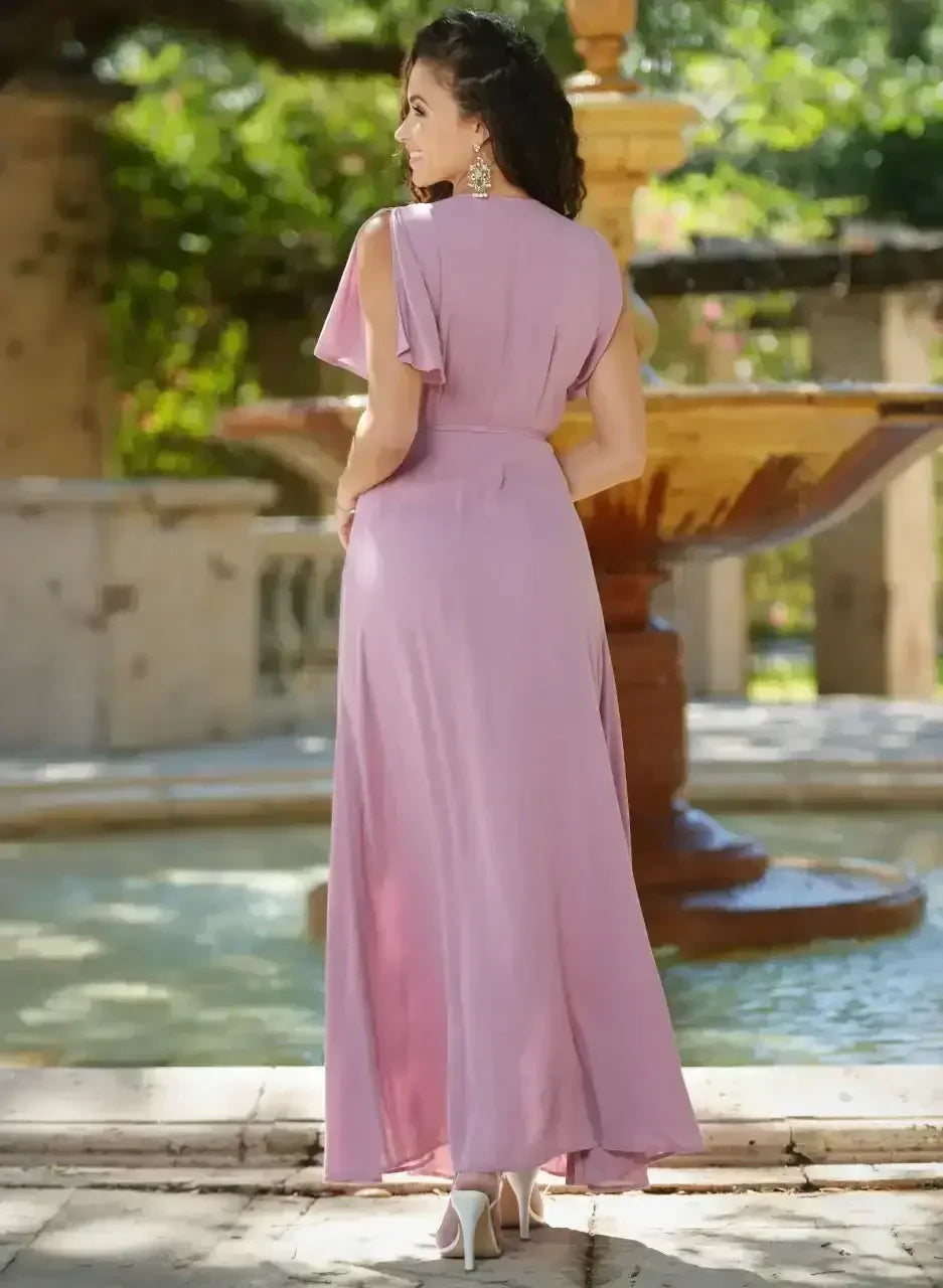 A woman in a flowing pink dress stands by a fountain, showcasing the dressâ€™s elegant silhouette. The lush greenery and sunlight create a serene and picturesque outdoor setting.