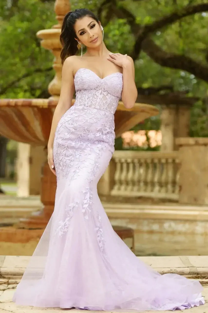 A woman in a strapless, lavender gown with floral details poses gracefully in an outdoor setting, featuring a fountain and lush greenery in the background.