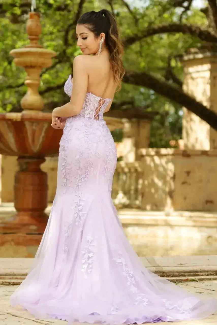 A young woman stands gracefully in a lavender gown adorned with floral details, with her back to the viewer, set against a lush garden backdrop featuring a fountain and stone structures.