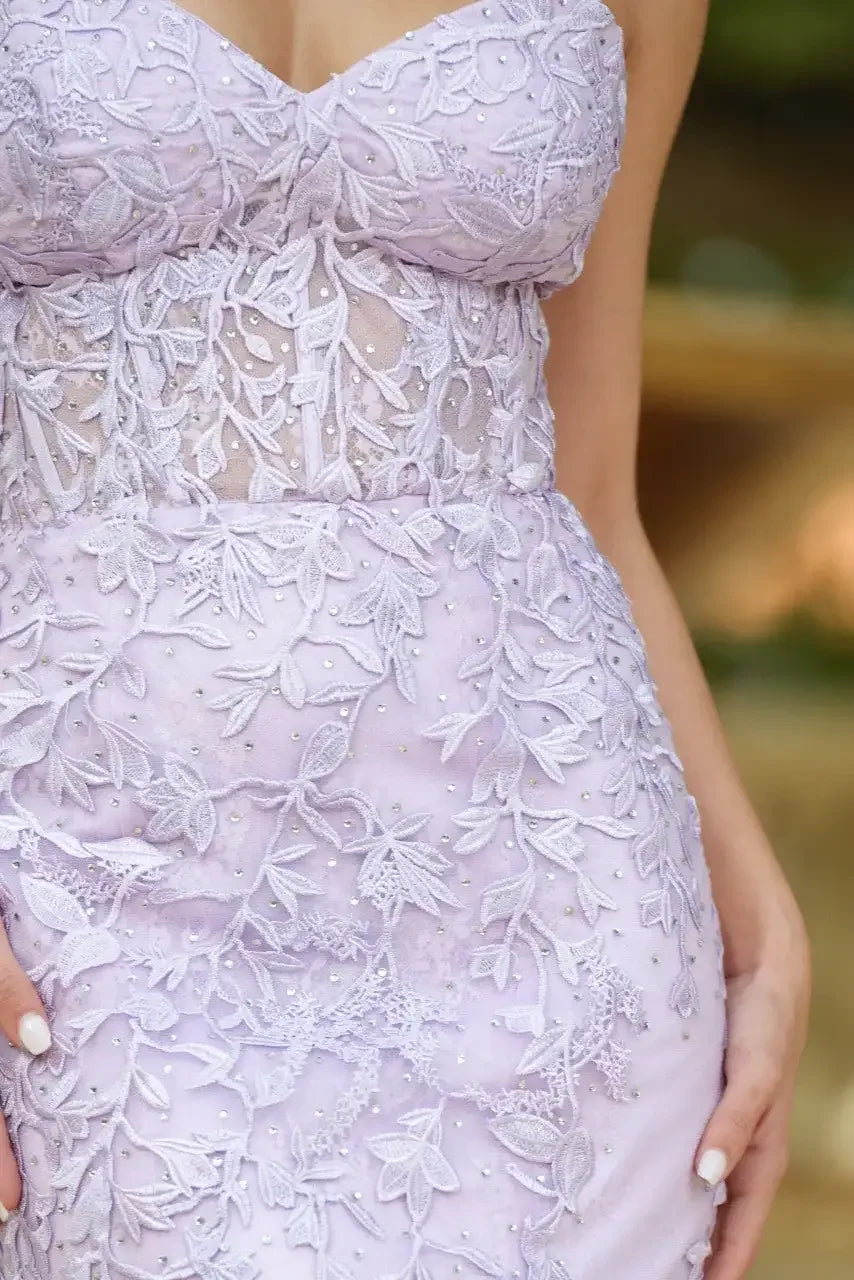 A close-up of a lilac dress adorned with intricate floral embroidery and embedded pearls. The person gently poses, showcasing the dress against a blurred natural background.