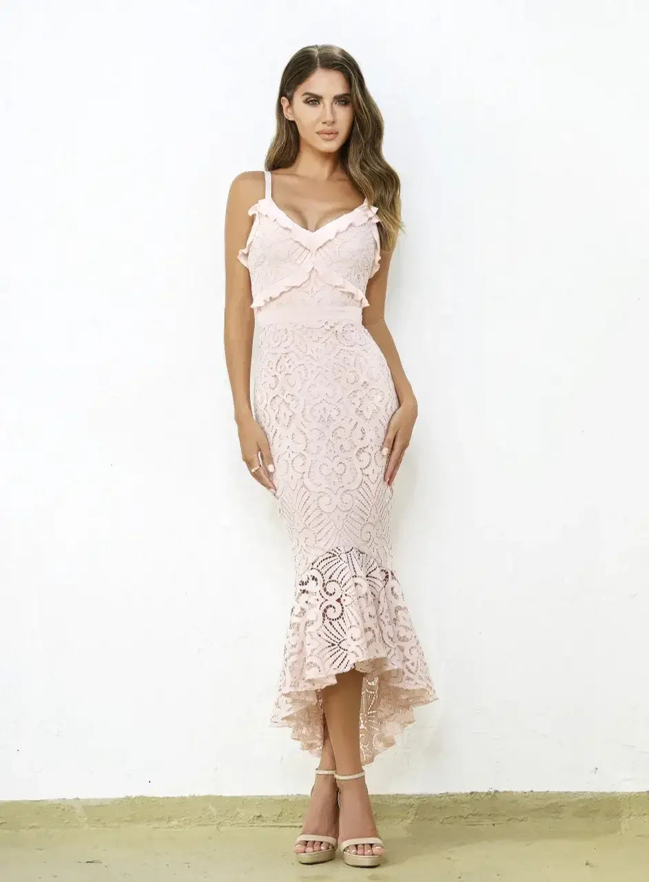 A woman stands confidently in a fitted, ankle-length, pink lace dress with ruffled straps. She is against a white wall with a beige floor, exuding elegance through her pose and styling.