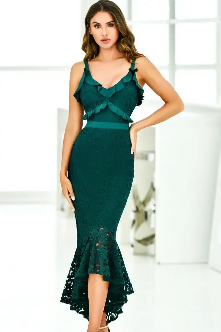A woman poses confidently in a form-fitting, dark green dress with ruffled straps and a flared hem. She stands in a bright, modern space with soft lighting and minimal decor.