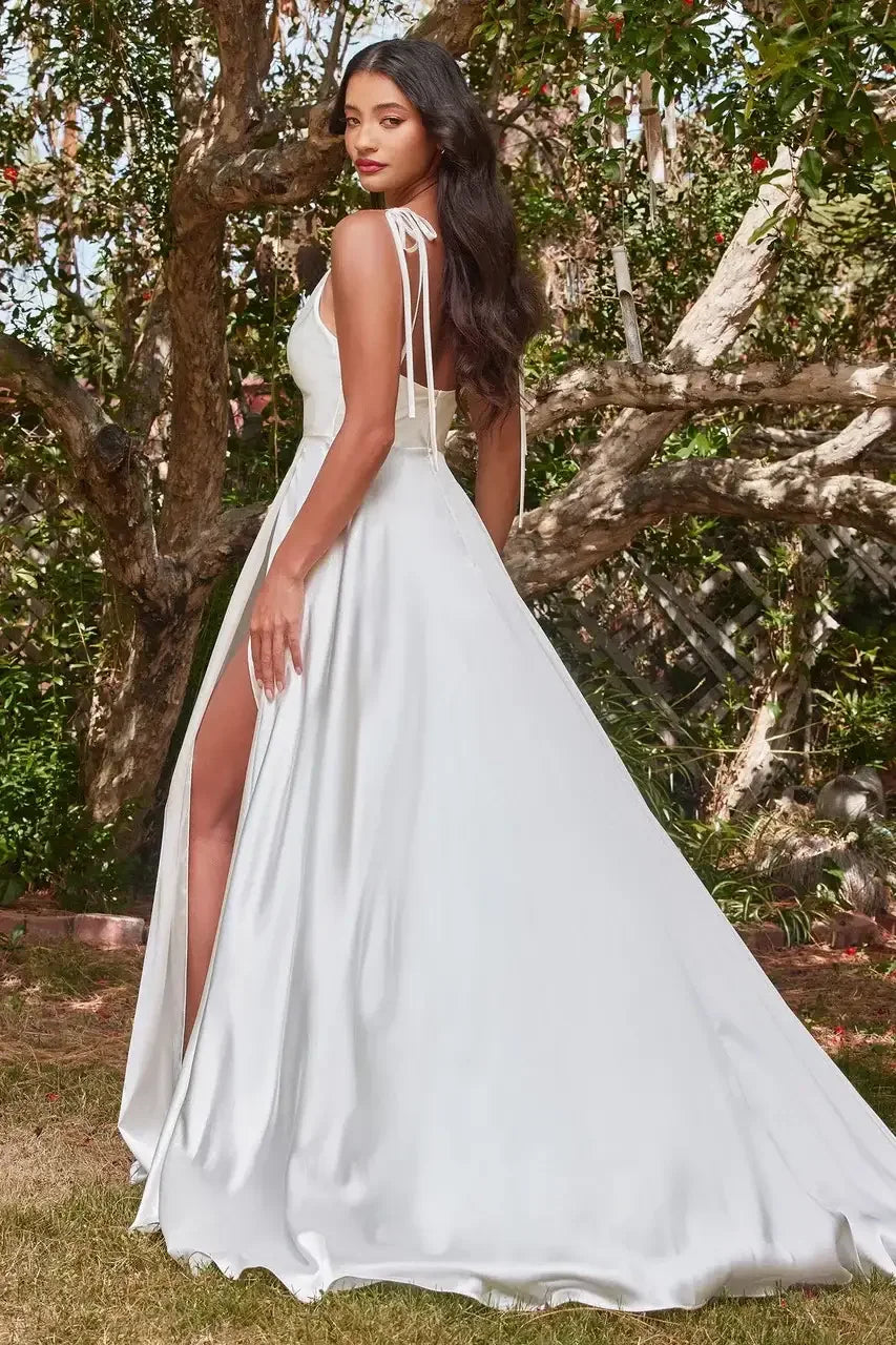 A woman in a flowing white gown stands elegantly, showcasing a high slit. She looks back over her shoulder, surrounded by greenery and trees in a natural setting.