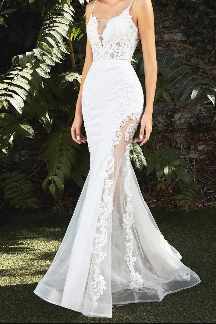 A woman wearing a form-fitting white gown with intricate lace details stands among lush green foliage. The dress features a sheer side, enhancing elegance and grace.