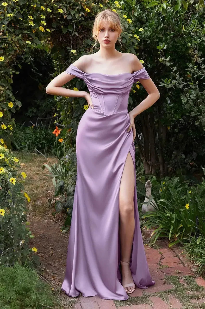A woman stands confidently in a lavender off-the-shoulder gown with a thigh-high slit, surrounded by vibrant flowers and greenery in a garden setting.