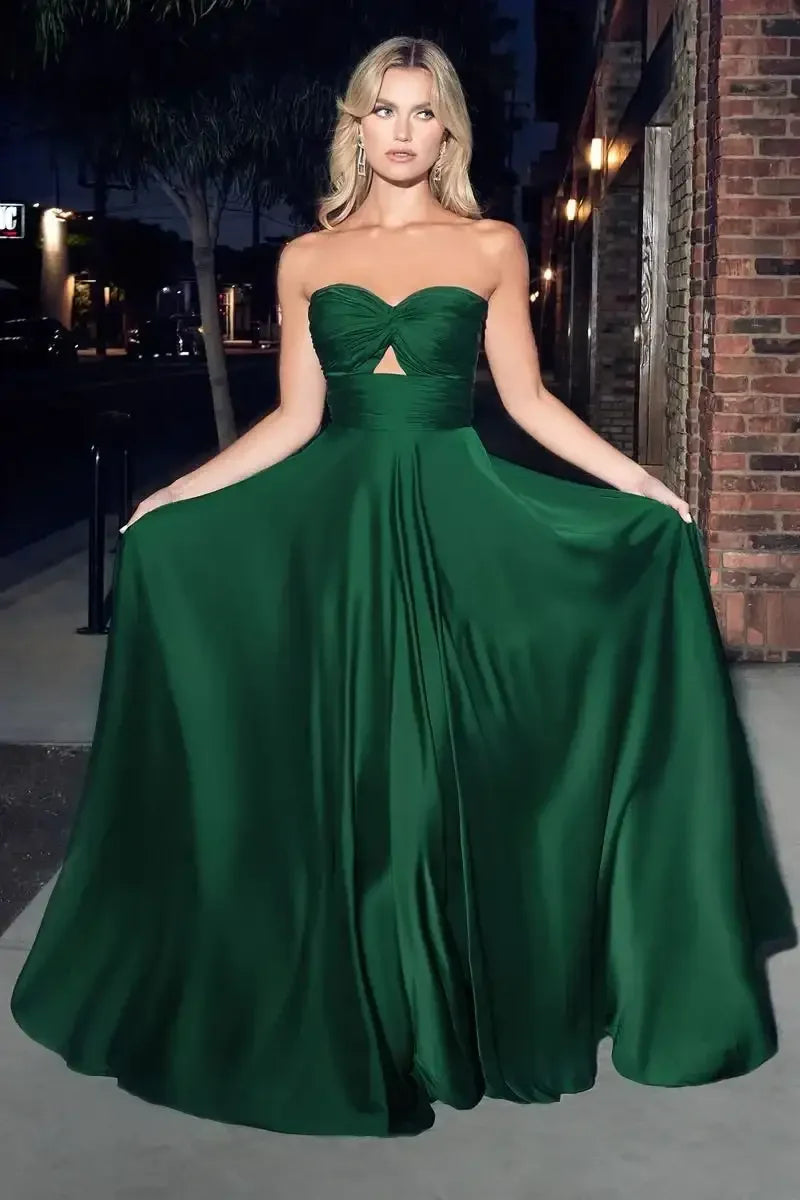 A woman in a strapless green gown stands gracefully, flowing fabric around her. She poses outdoors at night, with a softly lit urban background of buildings and trees.