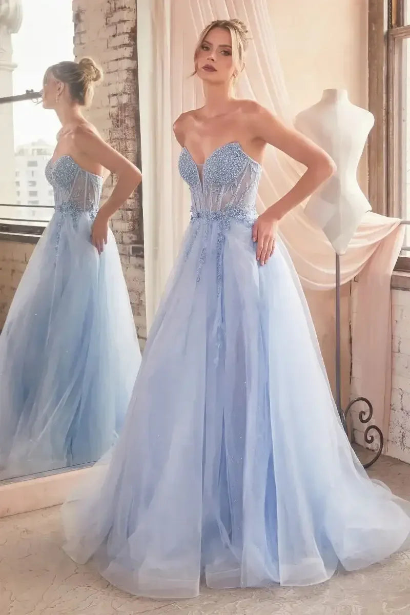 A woman poses in a light blue, strapless gown adorned with beads and tulle, reflecting in a mirror. Sunlight filters through a window, illuminating a vintage-styled room.