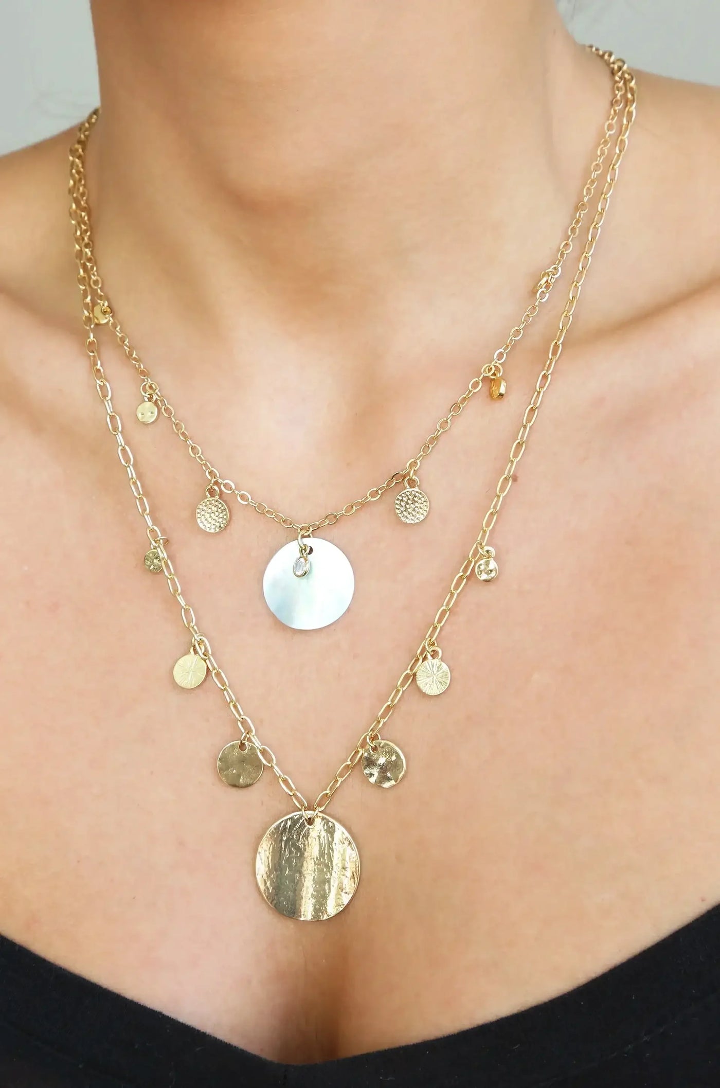 A layered gold and mother-of-pearl necklace rests elegantly on a person's neck, adorned with various circular charms. The wearer has a simple black top, with a neutral skin tone in a well-lit setting.