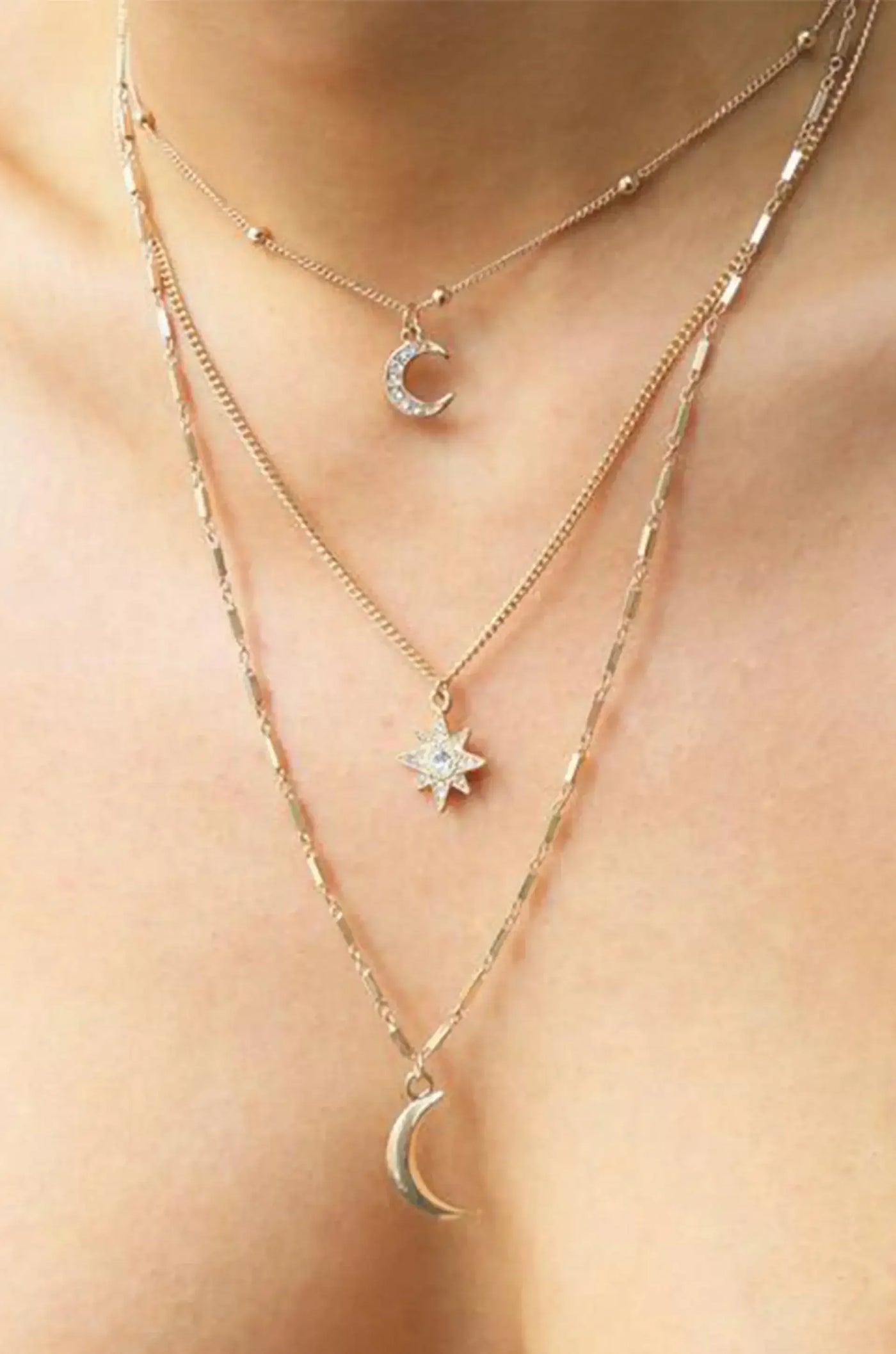 A layered gold necklace adorns a neck, featuring charms: a crescent moon, a star, and another crescent moon. The skin tone is visible, enhancing the jewelry's glow.