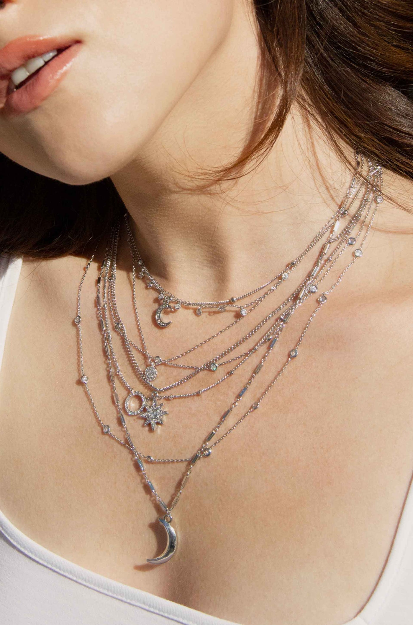 A series of silver necklaces drapes around a person's neck, featuring various pendants like a moon, star, and circular designs, against a soft background illuminated by natural light.
