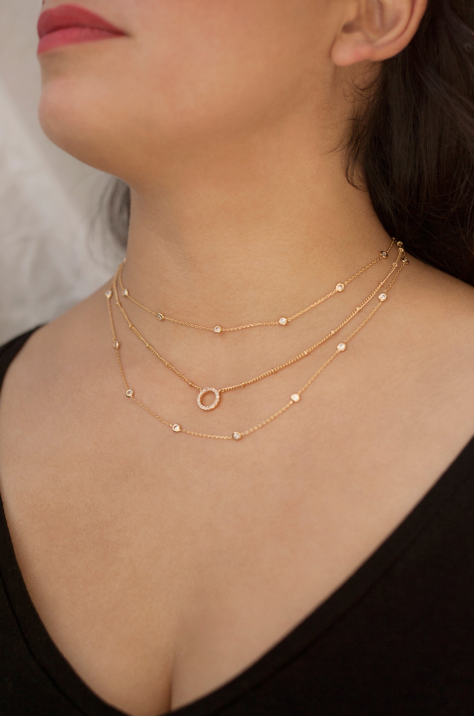 A triple-layered gold necklace with small beads and a circular pendant adorns a woman's neck, which is partially visible. She wears a black top, with soft lighting enhancing the scene.