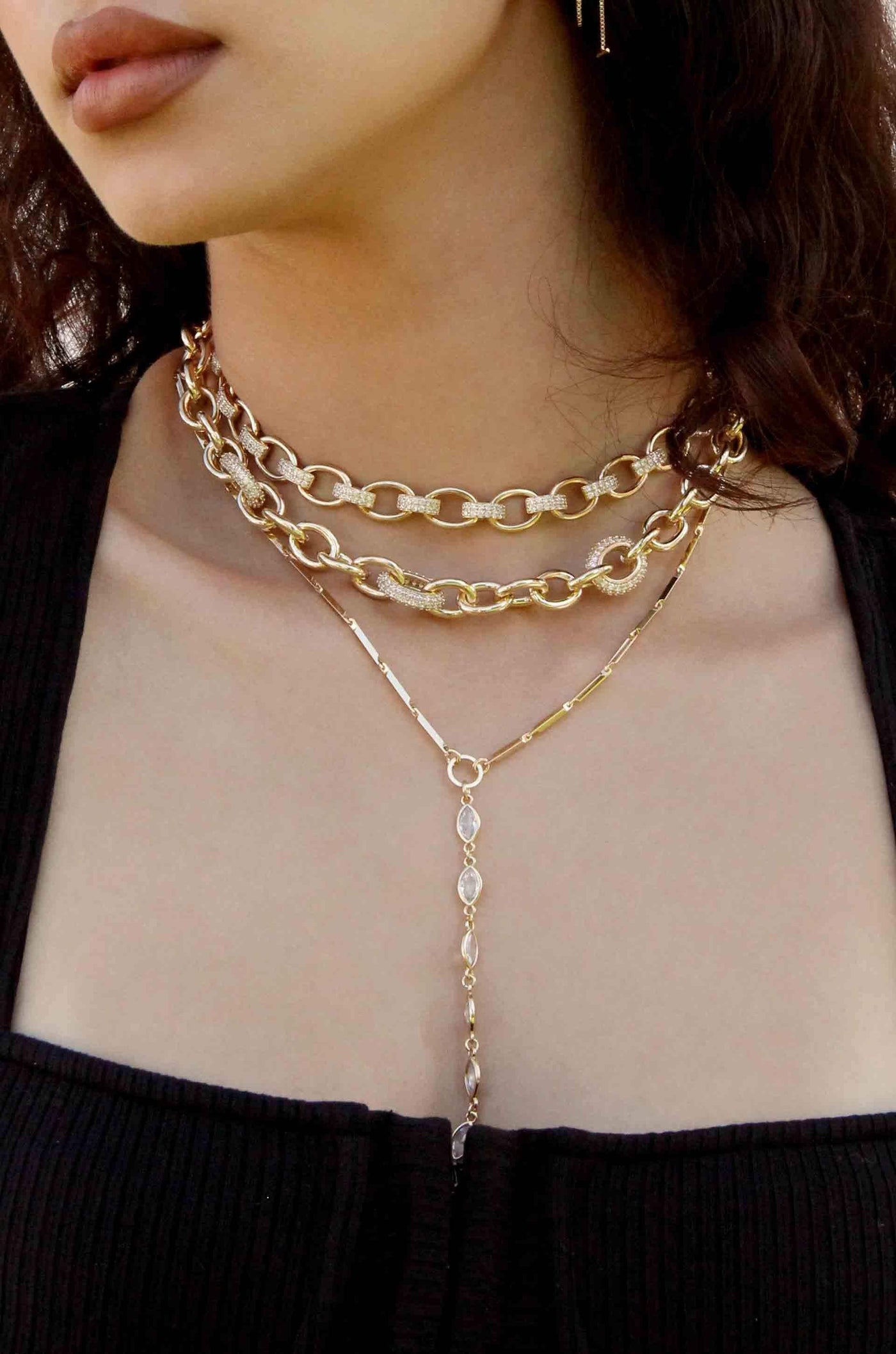 A layered gold necklace with multiple chains drapes elegantly around a person's neck. The jewelry contrasts with a dark, ribbed top and the skin tone, emphasizing the accessories' shine.