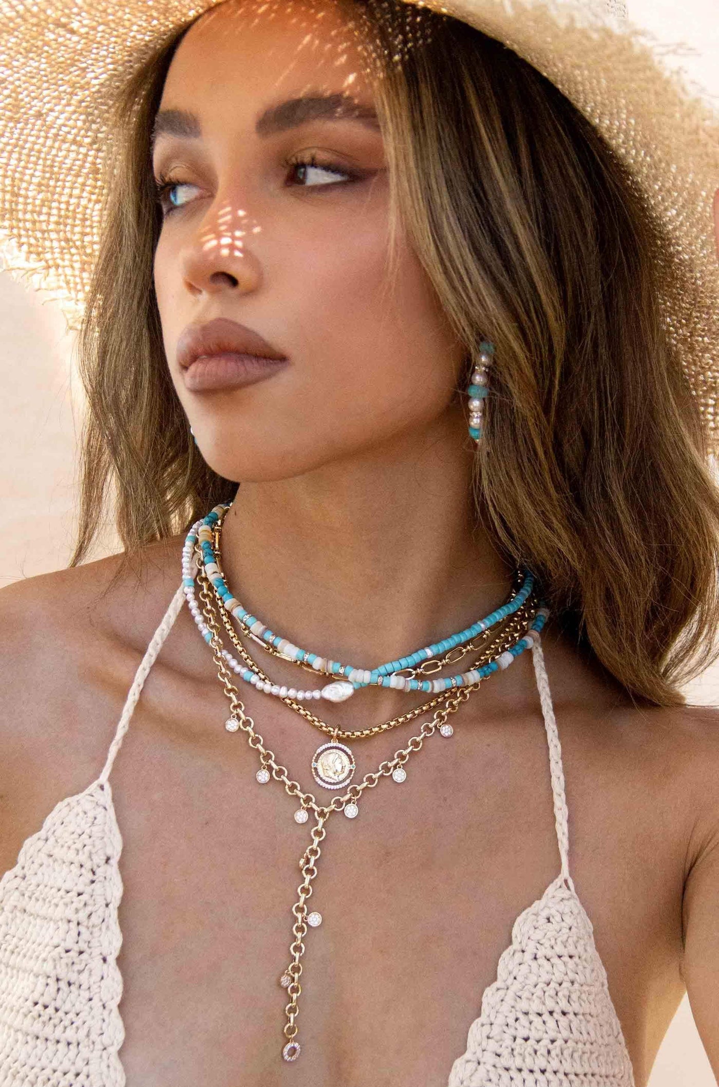 A woman wears layered necklaces featuring turquoise beads, pearls, and gold chains, set against a neutral background. Sunlight creates intricate patterns on her face and neck, enhancing the warm scene.