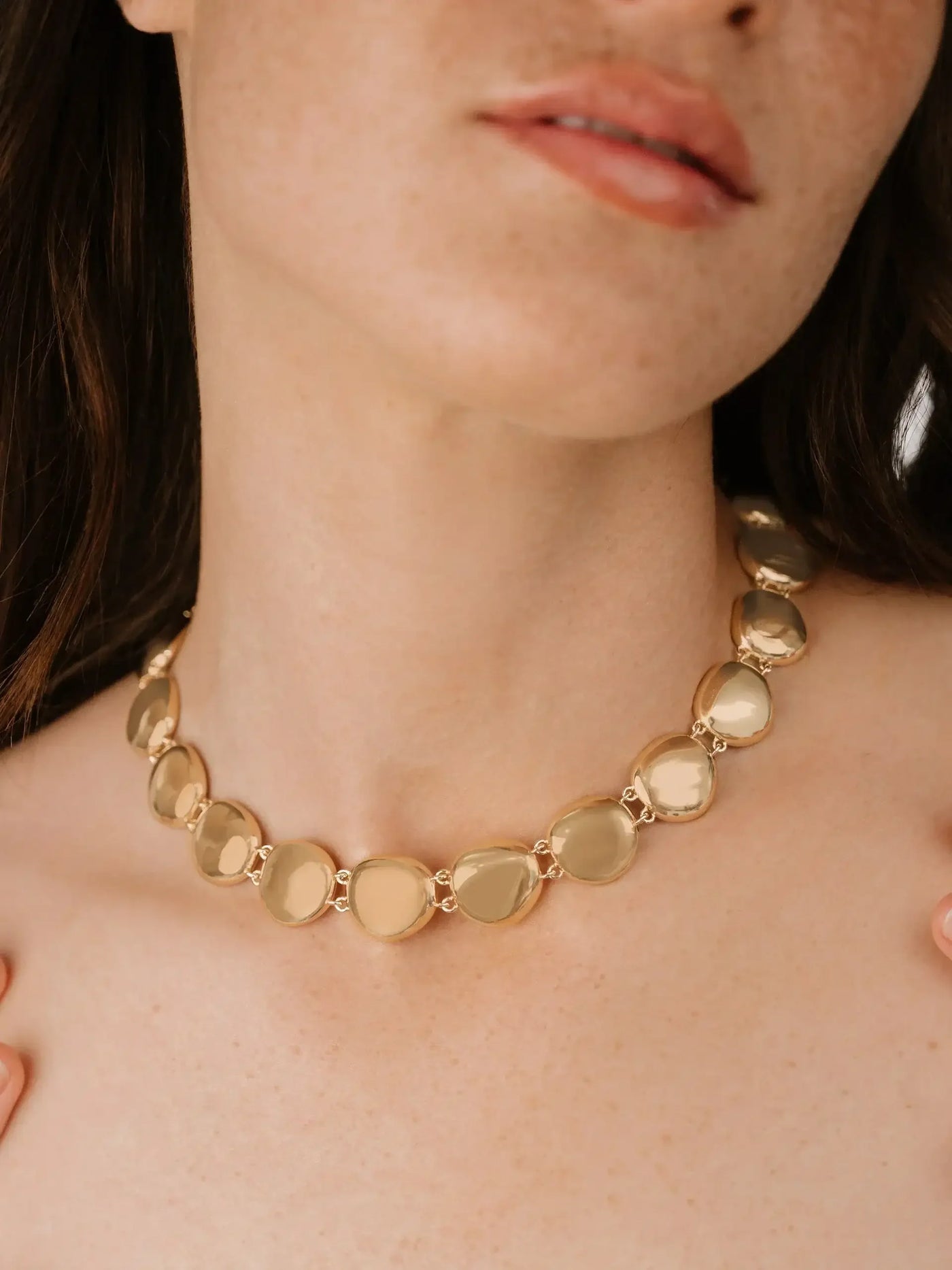 A gold-toned necklace with smooth, rounded stones rests on a woman's neck. She softly touches her collarbone while showcasing the jewelry against a neutral background.