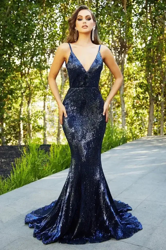 A woman stands confidently in a shimmering navy gown with a fitted bodice and flared skirt. She poses outdoors amidst lush greenery, emanating elegance and poise.