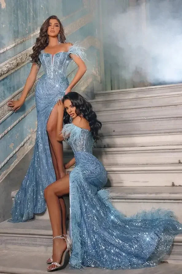 Two women in glamorous blue gowns pose on a staircase. One stands elegantly, while the other sits, highlighting their intricate outfits adorned with feathers and sequins, against a textured, vintage backdrop.