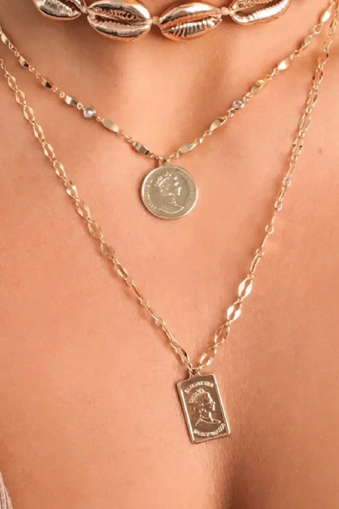 Gold necklaces with varying designs adorn a neck. The layered chains feature a round coin and a rectangular pendant, set against a softly lit skin background.