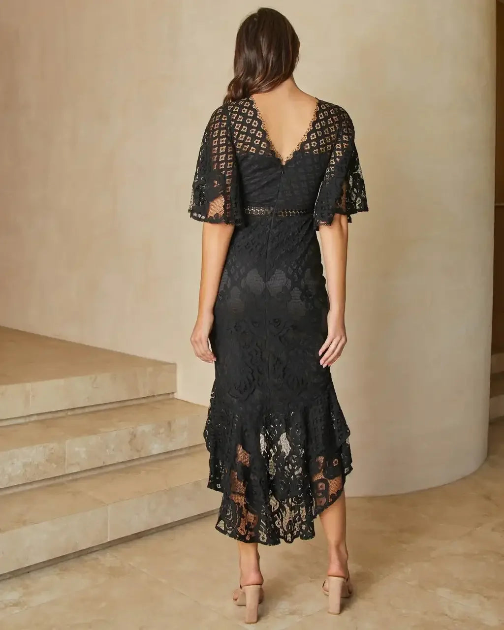 A woman wears a black lace dress with bell sleeves, standing on stairs in a minimalistic, light-colored interior, showcasing the intricate patterns of the fabric and a stylish, flowy hem.