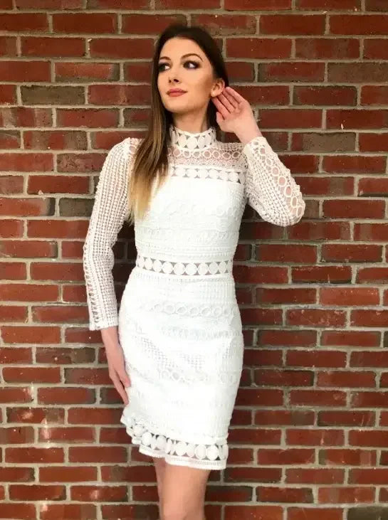A woman stands elegantly against a brick wall, wearing a stylish white lace dress with a high collar and intricate designs, holding her hair back while gazing thoughtfully.