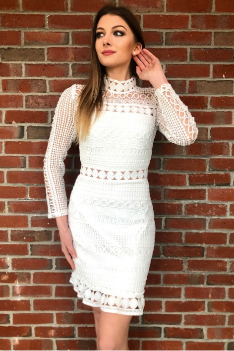 A woman stands elegantly against a brick wall, wearing a stylish white lace dress with a high collar and intricate designs, holding her hair back while gazing thoughtfully.
