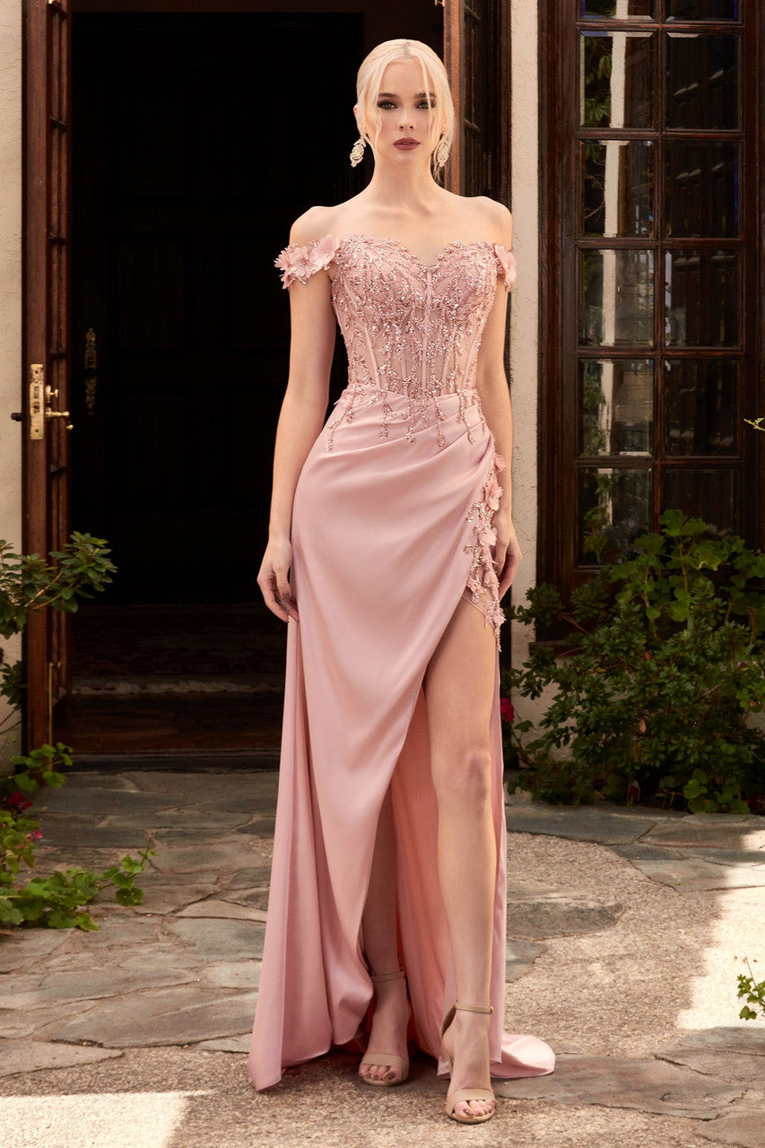 A woman stands elegantly in a pink gown with off-the-shoulder sleeves and intricate beadwork, showcasing a thigh-high slit. She is positioned in a landscaped outdoor setting by a doorway.