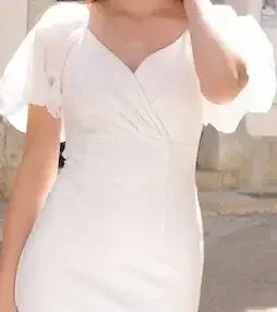 A person wearing a white dress with puff sleeves stands in a sunlit street. The dress features a v-neckline and a fitted bodice, set against a rustic backdrop.