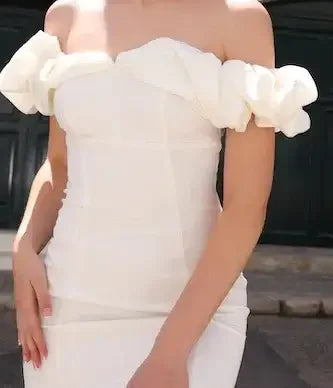 A white dress with an off-shoulder design features ruffled sleeves. A hand rests on the hip, highlighting the dress's fitted silhouette, against an outdoor background with a dark door.