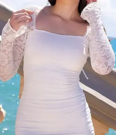 A person wears a fitted white dress with lace long sleeves, adjusting their outfit. The background features a bright outdoor setting near water, with blue skies and soft clouds.