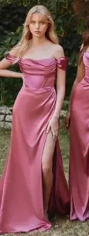 Two women stand side by side in elegant pink satin dresses with thigh-high slits. They pose in a lush outdoor setting with greenery in the background, showcasing a fashionable and sophisticated look.