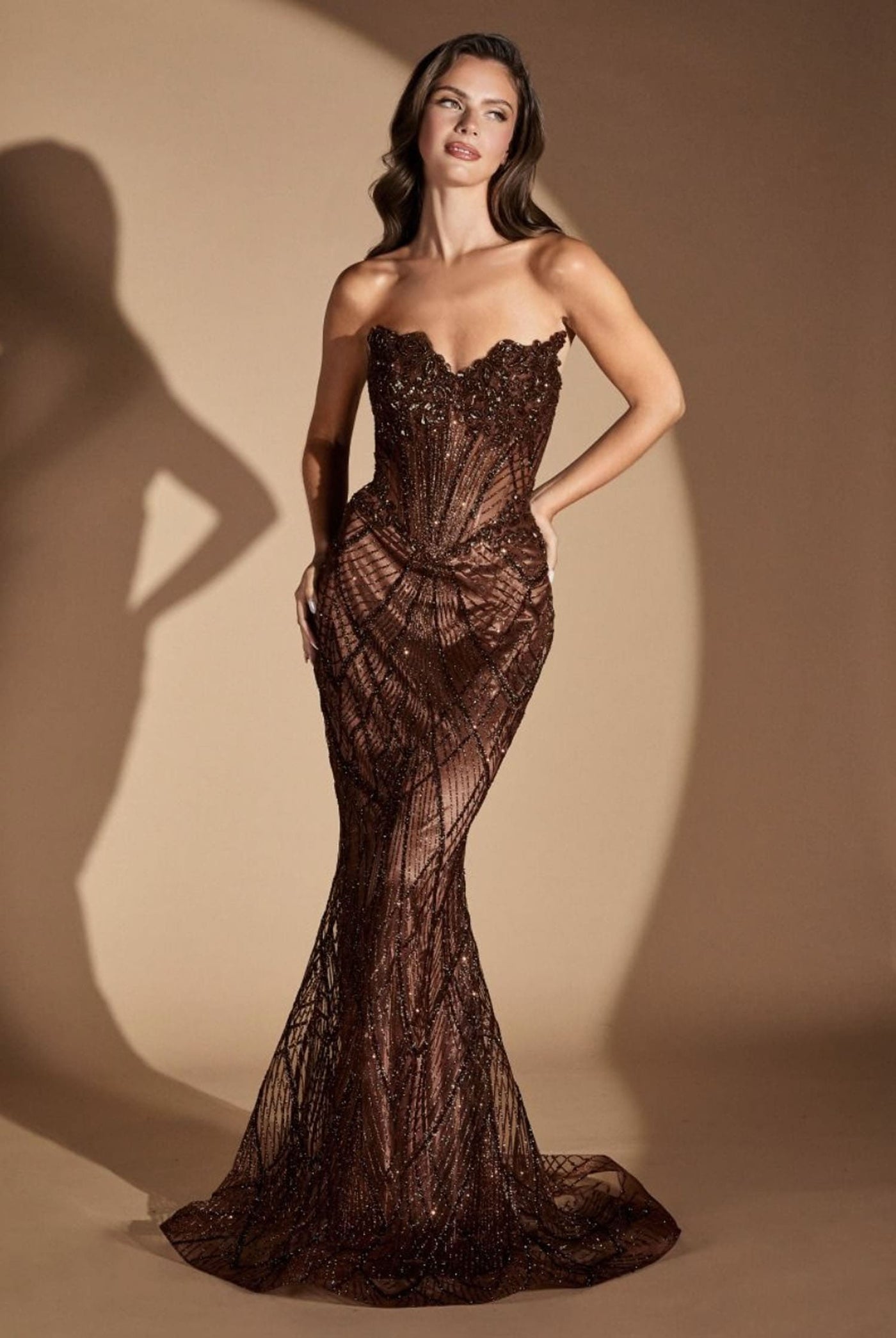 A woman models a strapless, sweetheart-neckline mermaid gown covered in dark brown sequins and beadwork, set against a warm beige studio backdrop with a soft shadow cast to the left.