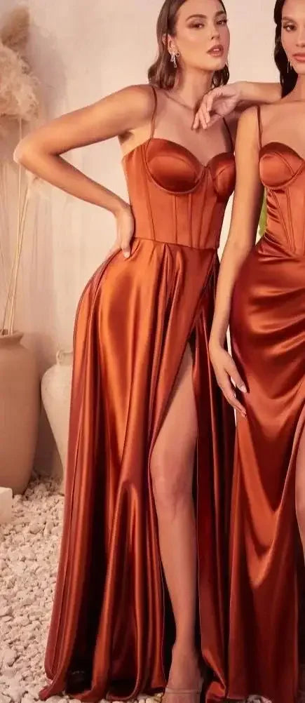 Two women are posing side by side, wearing long, satin, bronze dresses with thigh-high slits. They stand against a textured wall with wooden accents and a minimalistic background featuring stone and foliage.