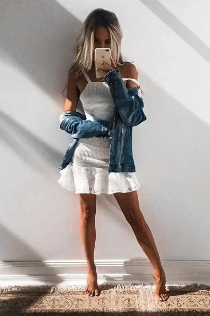 A woman stands barefoot, holding a phone, wearing a fitted white dress with a frilled hem and an oversized denim jacket, against a wall casting shadows and soft light.