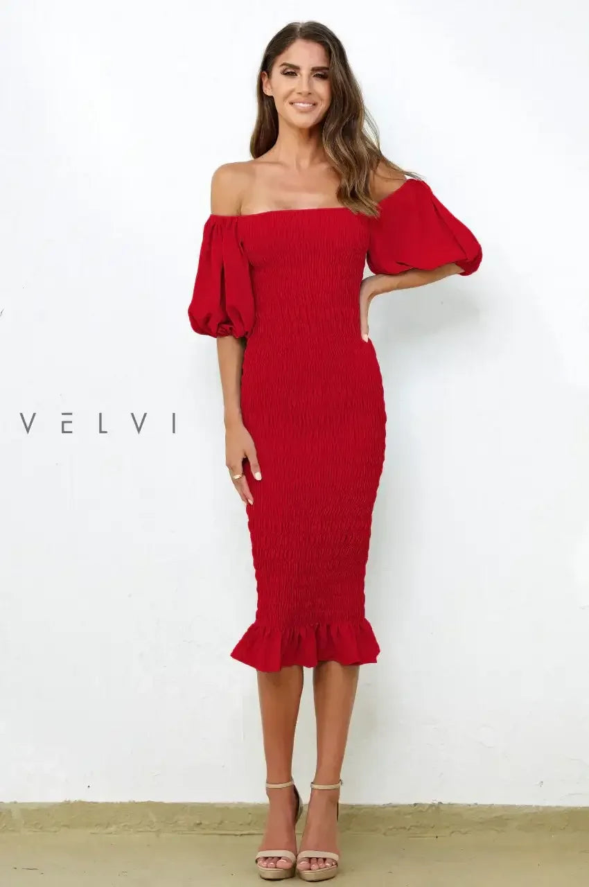 A woman wearing a fitted red dress with puffed sleeves poses confidently. The dress features a ruffled hem and a textured design, against a plain white wall in a neutral-toned setting.
