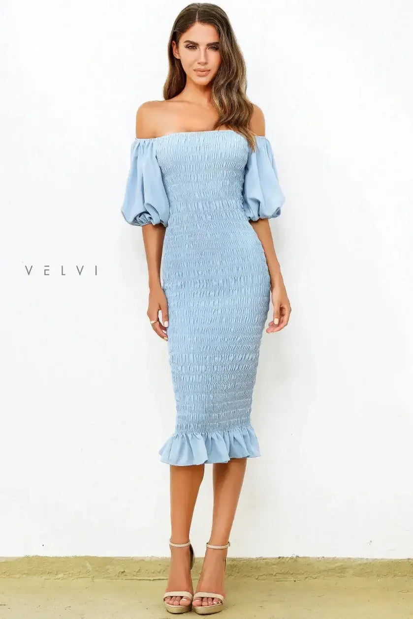 A woman stands confidently in a light blue, off-shoulder, ruched dress with puff sleeves and a ruffled hem. She wears strappy nude heels, set against a white wall backdrop.