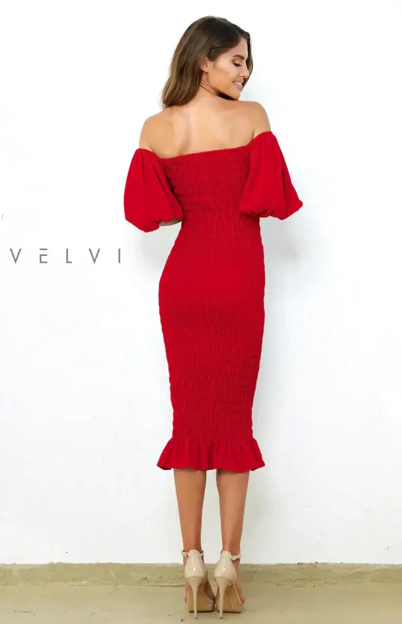 A woman stands with her back facing the viewer, wearing a fitted red off-the-shoulder dress with puffed sleeves, on a plain white wall background. She smiles gently, showcasing confidence.
