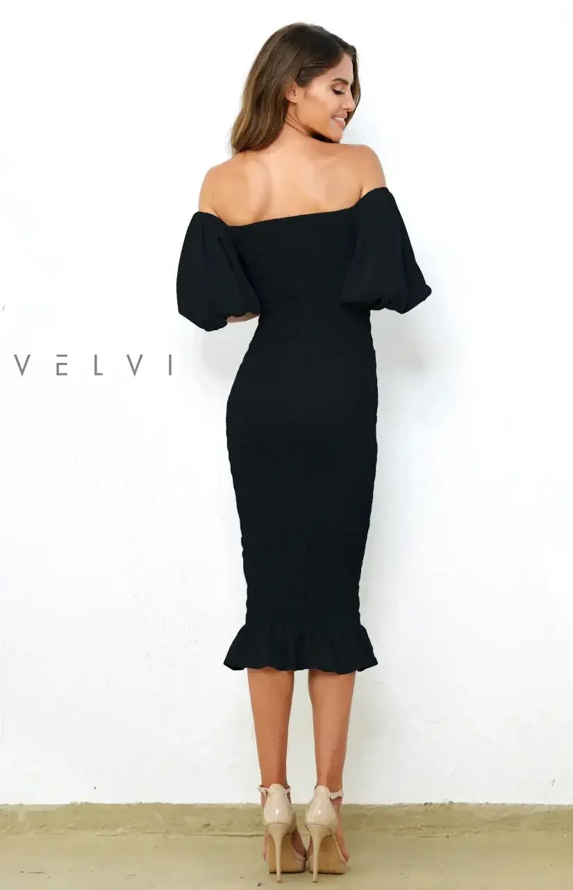 A woman stands with her back to the viewer, wearing a fitted black dress with puff sleeves and a ruffled hem. She has wavy hair and is in front of a white wall.