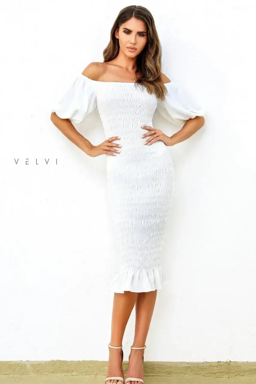 A woman wears a fitted off-shoulder white dress with puff sleeves and a ruffled hem, standing against a plain white wall, posing with arms crossed confidently.