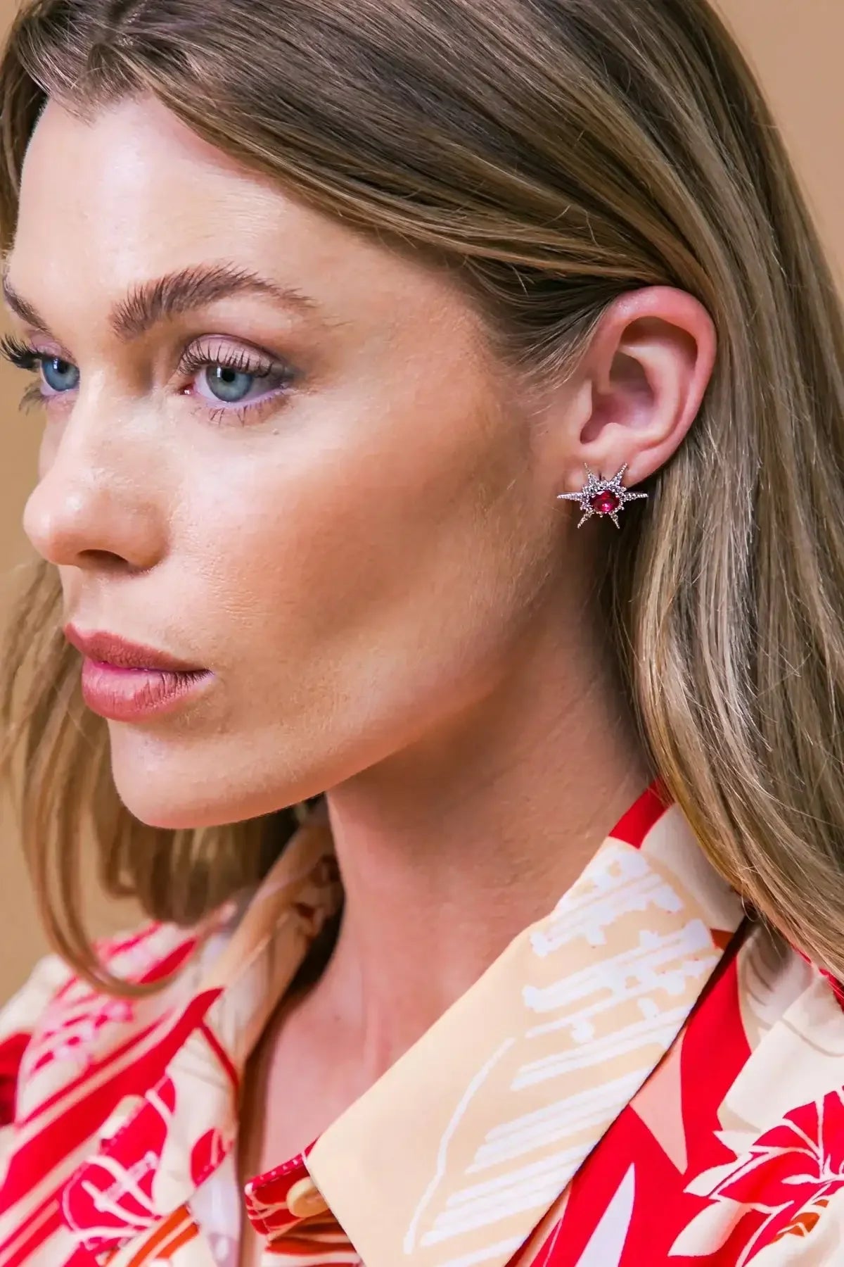 A woman with light skin and long, wavy hair wears a striking star-shaped earring featuring a red gemstone. She's dressed in a colorful, patterned blouse, set against a neutral background.