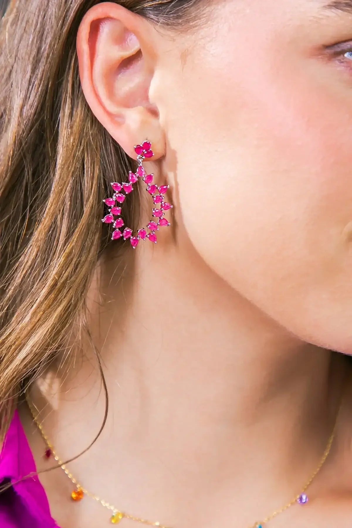 A close-up of a model's ear features a large, striking pink earring adorned with various shaped stones, while the model has a subtle smile and wears a gold necklace with colorful accents.