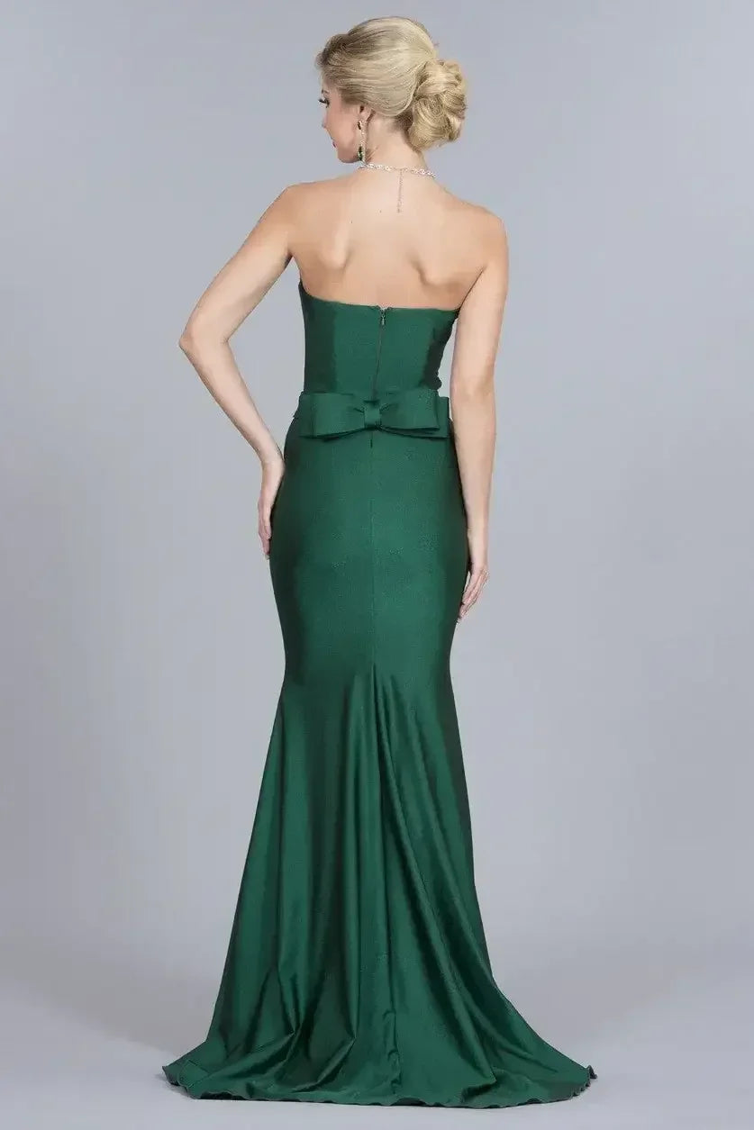 A woman in a strapless green gown stands gracefully with her back to the viewer, showcasing the fitted silhouette and a large bow detail at the waist against a neutral background.
