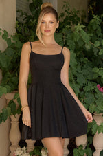 A woman wearing a black spaghetti-strap mini dress with a flared skirt, standing outdoors in front of green potted plants and stone balustrades.