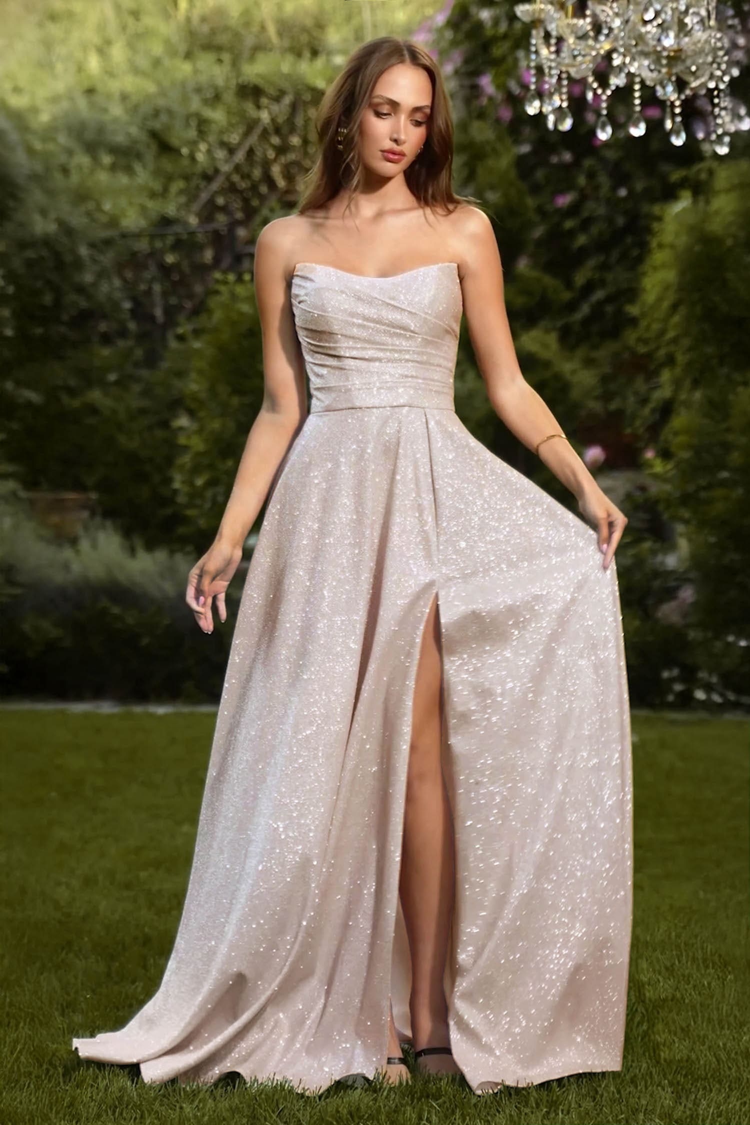 A woman stands outdoors on grass wearing a strapless floor-length gown covered in shimmering sequins. The dress has a fitted bodice, a high front slit, and a flowing skirt.